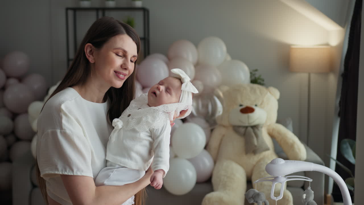 Mãe abraça ternamente sua recém-nascida cercada por balões festivos e um grande urso de pelúcia.