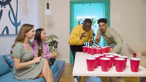 happy multiethnic young friends playing beer pong at home