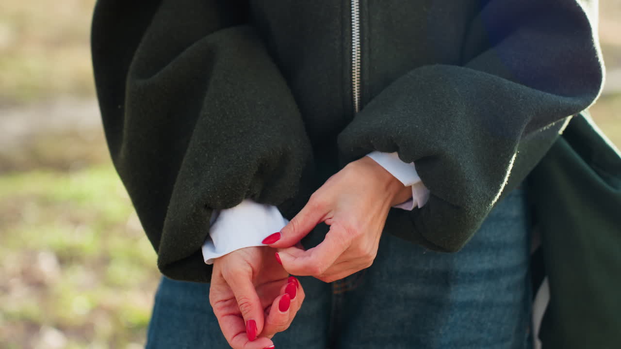 Closeup of woman with red manicure adjusting hand near sleeve of dark jacket and white shirt, standing outdoors on blurred green natural background with soft daylight focus