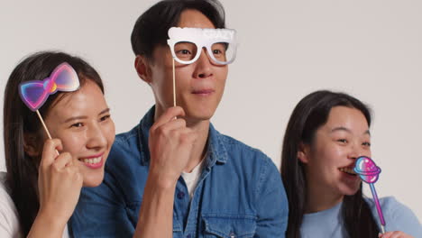 Side-View-Of-Group-Of-Young-Friends-In-Front-Of-White-Studio-Background-Having-Fun-Posing-For-Photo-Booth-Style-Portraits-With-Props-1