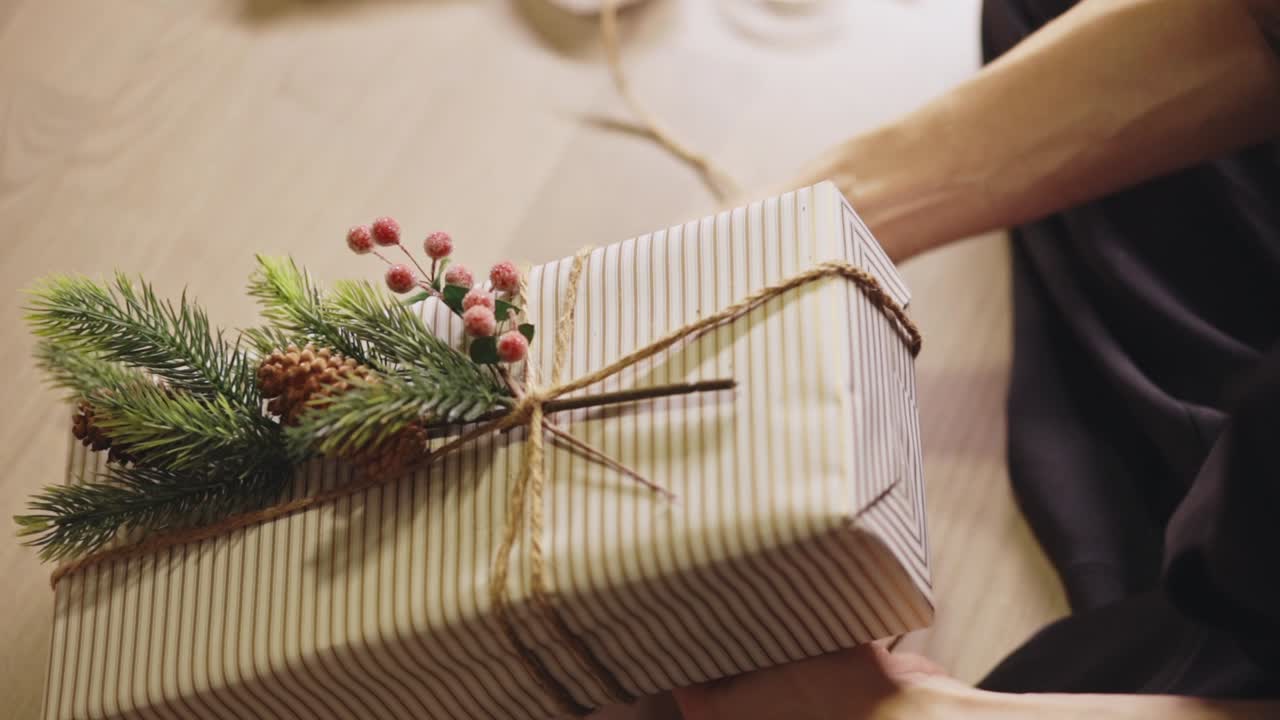 Hands Wrap Twine Around a Striped Gift Topped With Pine Sprigs, Adding a Festive Christmas Touch - Close Up