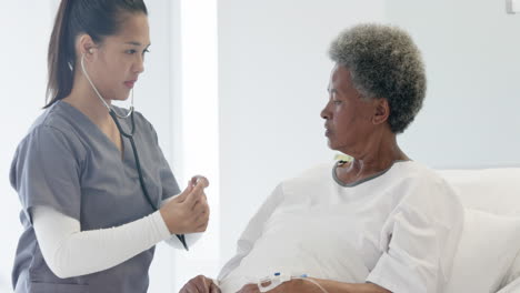 diverse female doctor testing senior female patient using stethoscope in hospital room, slow motion