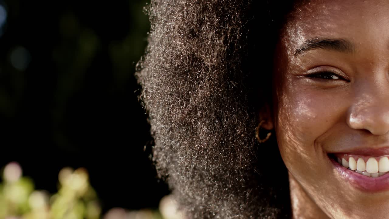 Donna afroamericana che sorride ampiamente in un giardino illuminato dal sole, con i capelli e un orecchino a cerchio d'oro in evidenza.