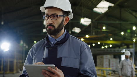 trabajador de una fábrica caucásica con casco y gafas, escribiendo en una tableta en una gran fábrica