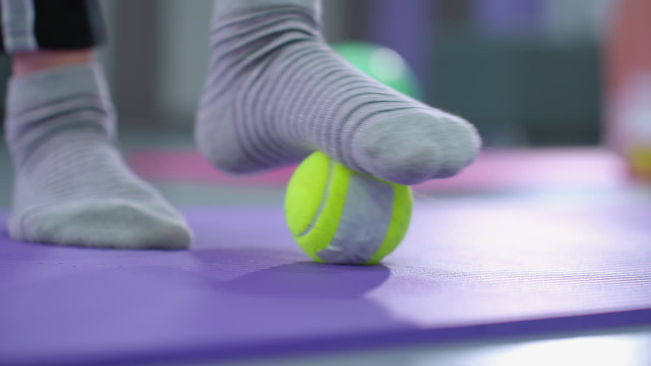 양말을 입고 보라색 매트 위에 노란색 테니스 공을 굴리는 사람의 발의 클로즈업, 부드러운 흐릿한 배경과 빛의 반사, 발 치료에 초점을 맞추고