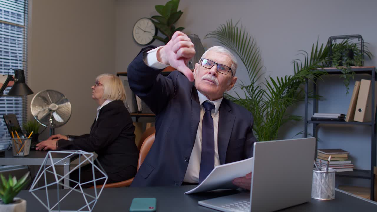 Unhappy elderly man boss making thumb down gesture expressing discontent, disapproval in office room