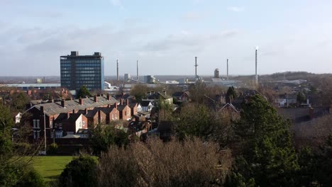 Aerial-zoom-in-view-over-park-trees-to-industrial-townscape-with-blue-skyscraper,-Merseyside,-England