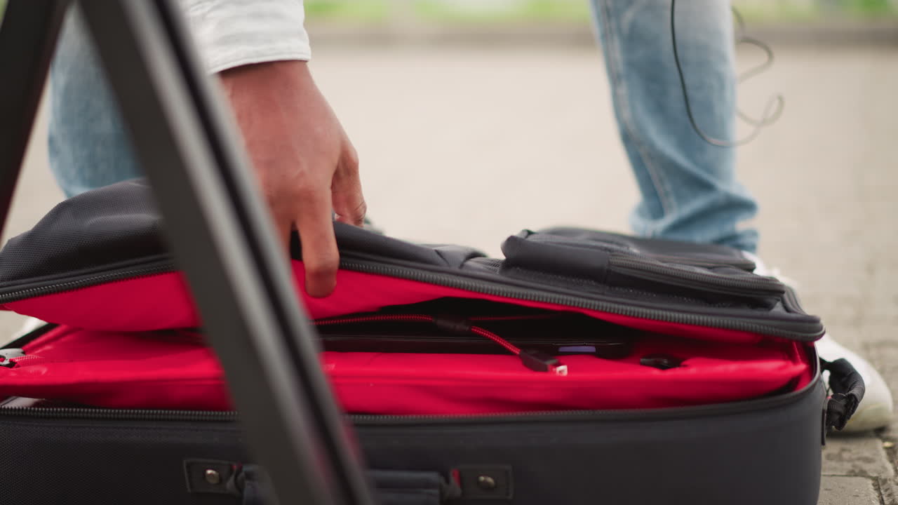 Mala com forro vermelho no pavimento, músico verifica cabos e bolsa enquanto ajusta o tripé, close-up de pés e mão em jeans prendendo carregador e cabos de áudio, preparação urbana tensa e focada.