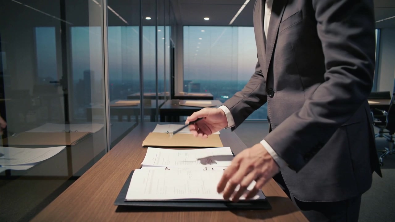 Un uomo in abito scuro firma documenti, esaminando un raccoglitore e una cartella di cartone al tavolo dell'ufficio, mentre cala il crepuscolo.