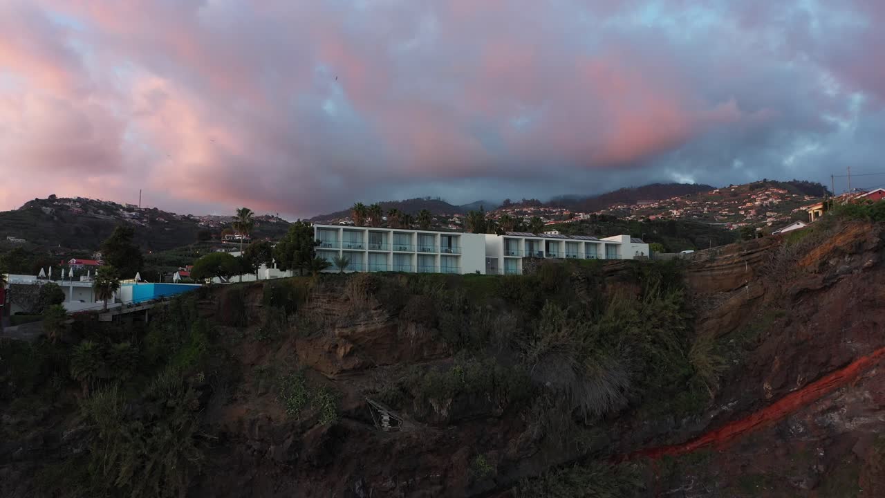 drone disparado moviéndose hacia arriba y inclinándose hacia abajo en el hotel de lujo en el acantilado de ponta do sol en madeira durante la puesta de sol con dramáticas nubes de tormenta en el fondo