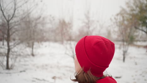 close-up of woman wearing red beanie and hoodie jogging outdoors on a snowy winter day, surrounded by serene park trees and evergreen pines, with blurred background