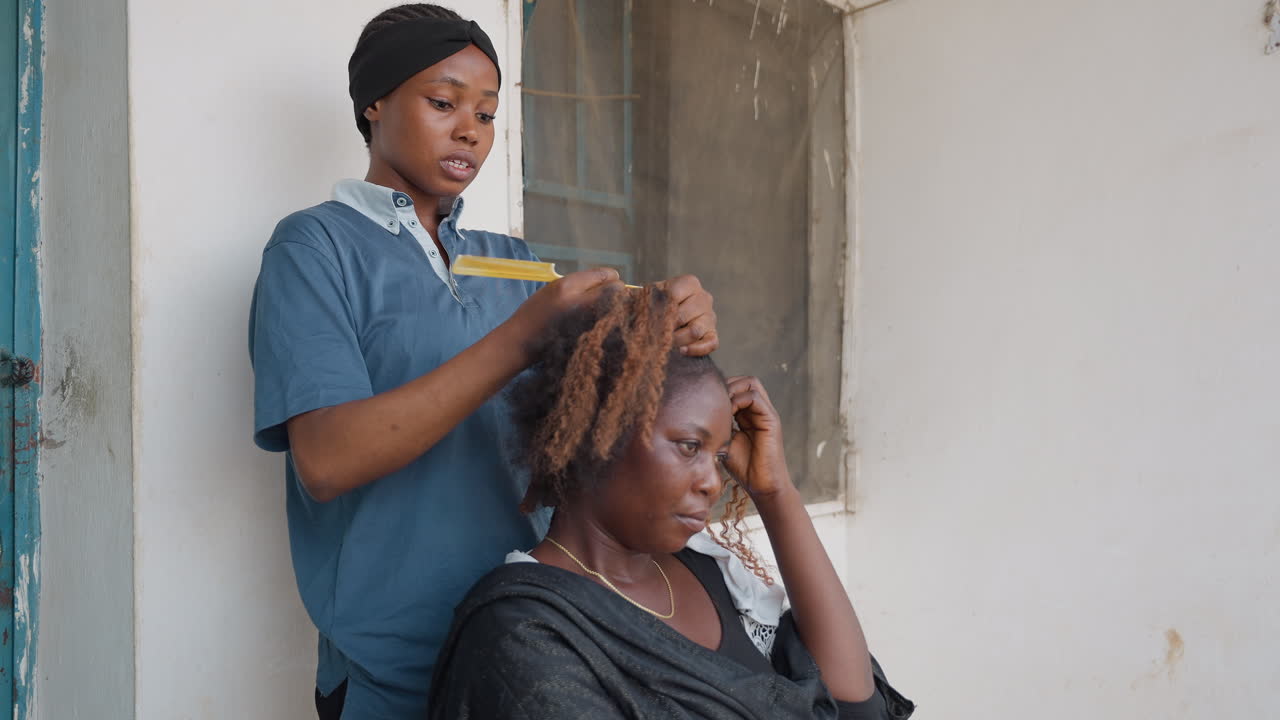 Stilista nero che intreccia i capelli del cliente, apprendista in piedi che pettina ricci testurizzati su cliente seduto, espressione concentrata, mani che lavorano con il pettine, ambiente casuale all'ingresso, scena di styling collaborativa.