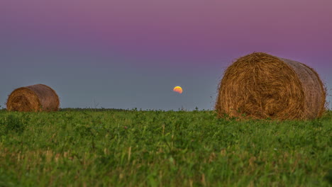 Moon-setting-down-behind-green-meadow-and-haybales,-time-lapse-view
