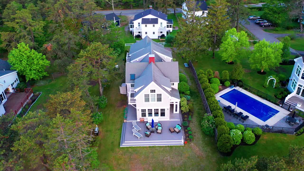 Stunning drone real estate video of a lovely suburban cottage house with a wide back porch, stylish patio furniture, and a well-kept backyard, all surrounded by tall green trees and blooming shrubs