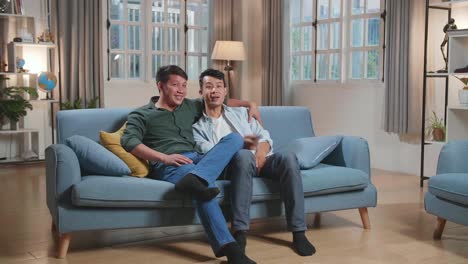 happy asian gay couple watching tv, while sitting on a couch in cozy stylish apartment. adult boyfriends hug, watching funny videos and streaming movies from online service