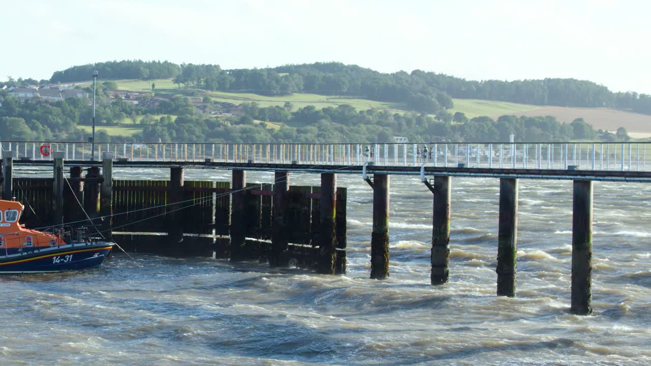 Choppy sea waves hit a pier with lifeboat, hills and countryside visible in background