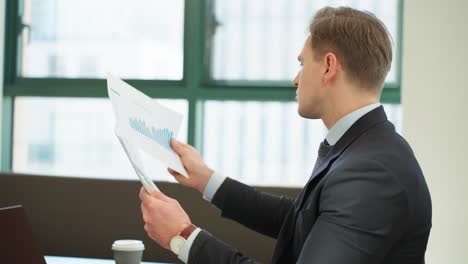 caucasian professional man analyzing printed charts graphs, nodes with acceptance and approval, happy with successful results, drinks coffee in a well-lit office with large windows - parallax midshot