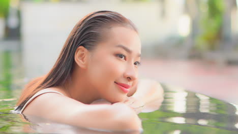 face close-up of asian woman with perfect skin leaning on arms and smiling on edge of swimming pool at topical resort in bali