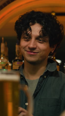 Vertical-shot-of-Attractive-young-bartender-stands-behind-the-bar-counter,-smiles-and-communicates-with-visitors.-Mature-man-talks-with-friend,-spends-weekend-in-stylish-pub-at-night.-Concept-of-lifestyle-and-leisure.