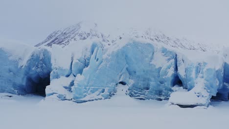 Front-of-glacier-with-blue-ice-and-multiple-caves,-mountains-as-backdrop,-aerial