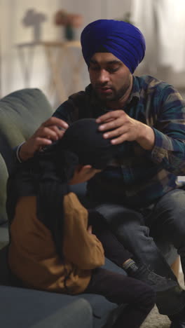 vertical video shot of father tying turban onto head of young sikh son with top knot sitting on sofa at home