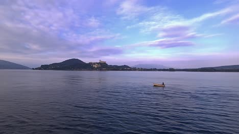 wide angle view of small fishing boat with fisherman rowing in calm lake waters of maggiore lake in italy with angera castle in background
