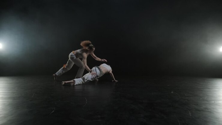 Gruppo di ballerini professionisti che praticano capoeira nell'oscurità contro un riflettore su uno sfondo nero dello studio. Arte marziale afro-brasiliana che combina elementi di danza