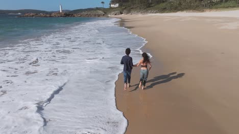 couple running in the shore of beach with waves. - aerial follow shot