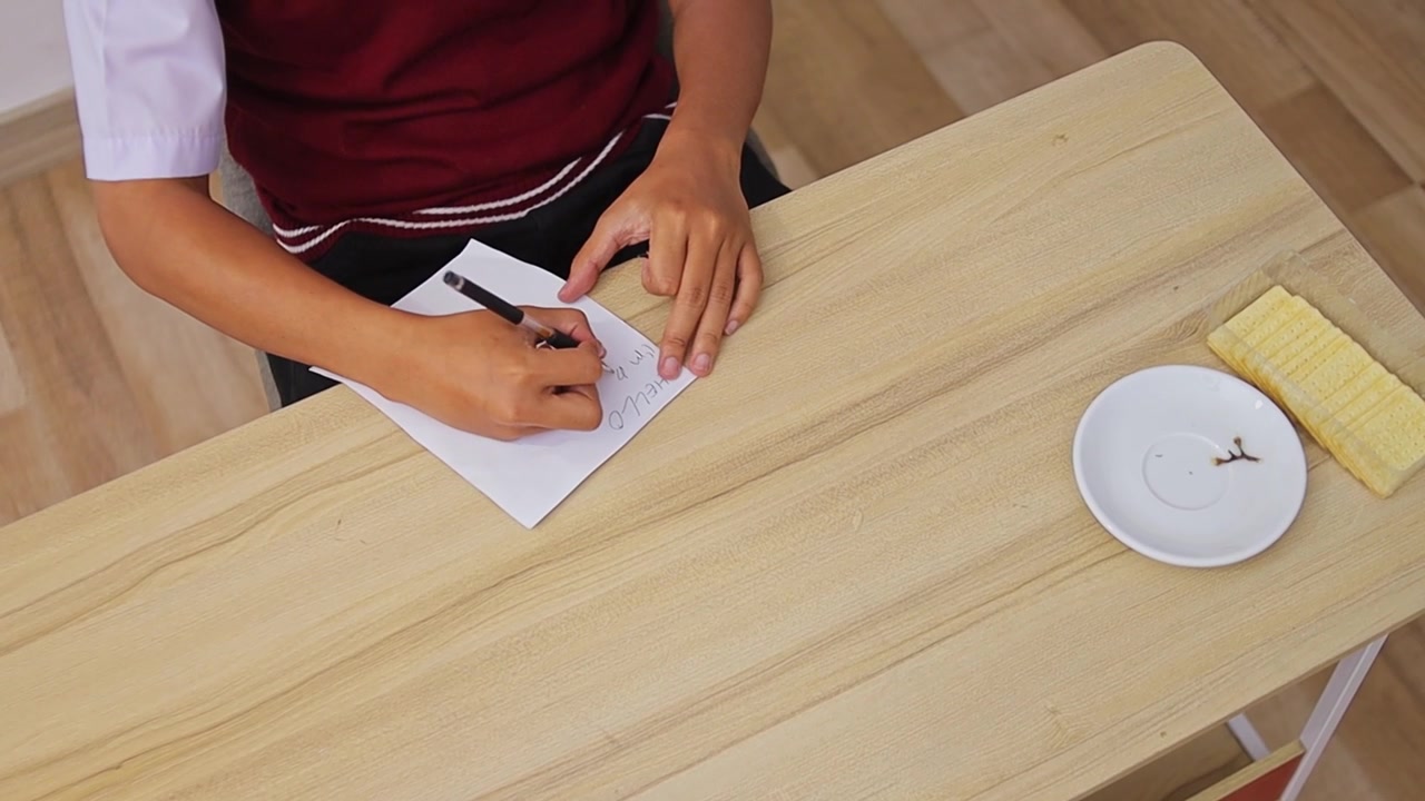 Overhead view of student in uniform writing "Hello" greeting on sheet of paper at wooden desk