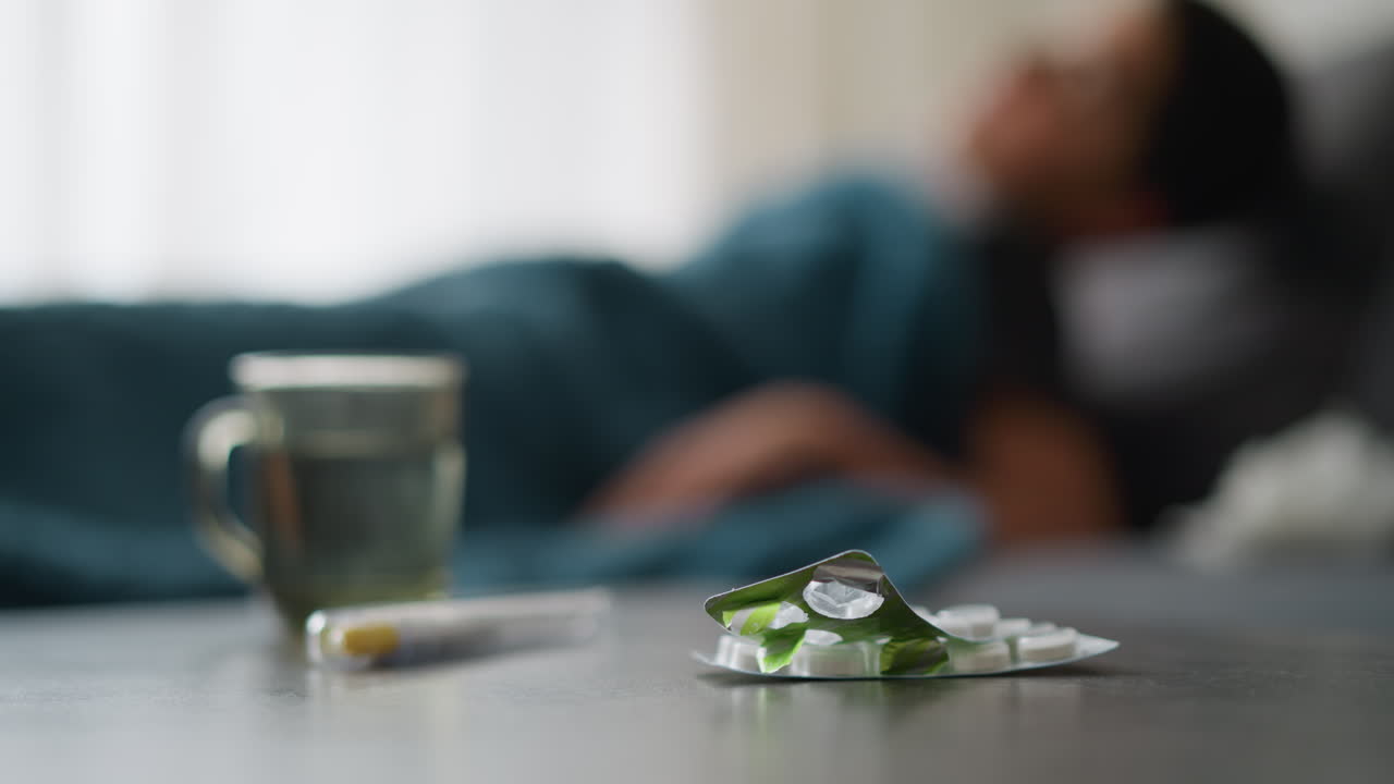 Close-up of medicine and cup on table with blurred view of someone adjusting on bed, relaxing, peaceful setting, possibly indicating a sick day or recovery period