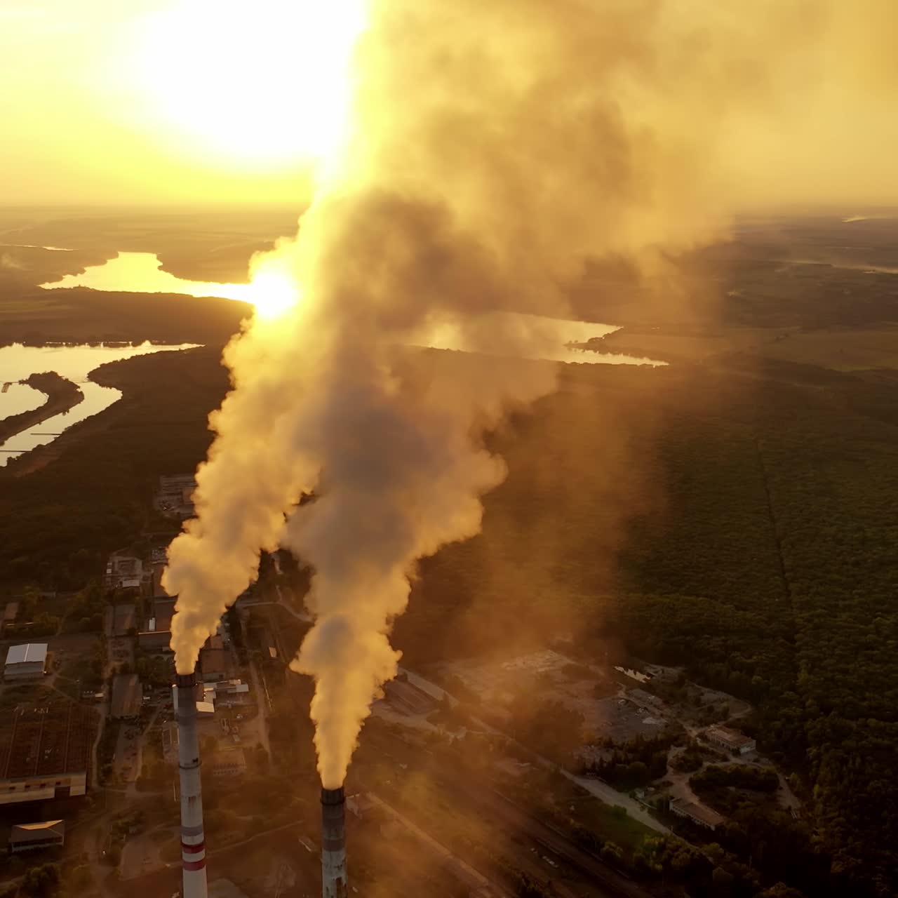 Industrial factory near the river. Top view of thick smoke coming from pipes into the air at sunset. Manufacturing among nature. Aerial view.