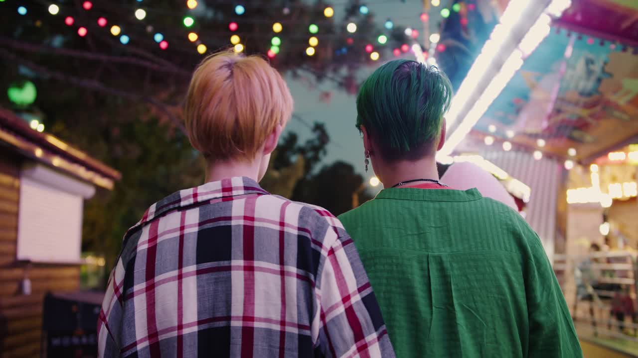 Rear view of a blonde girl with a short haircut in a plaid shirt and a girl with a green short haircut in a green shirt walk through a bright and glowing amusement park during their date