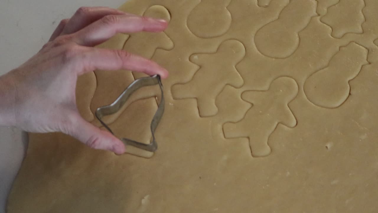 Un primo piano che ritaglia varie forme di biscotti natalizi dalla pasta frolla. Accanto a biscotti a forma di omino di pan di zenzero e pupazzo di neve, viene utilizzato un tagliabiscotti a forma di campana.