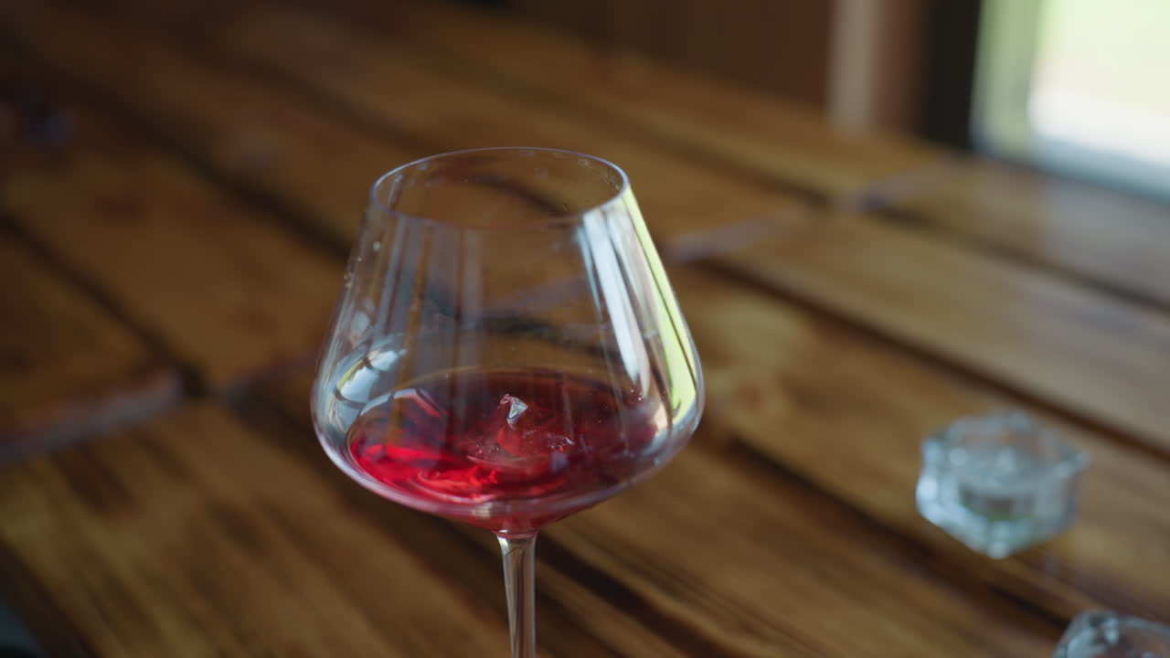 closeup of hand gently dropping ice cube into wine glass filled with red drink on polished wooden table with natural daylight streaming through window creating warm ambient indoor setting