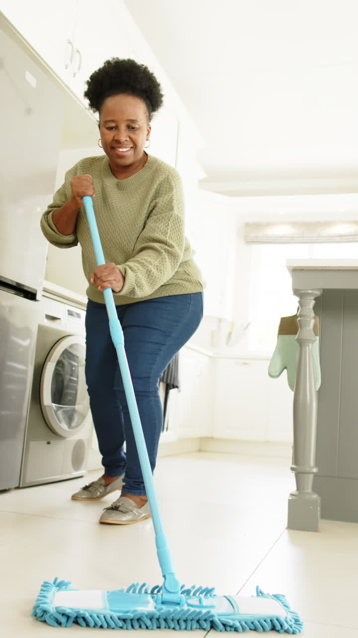 Happy african american senior woman cleaning floor in sunny kitchen, slow motion