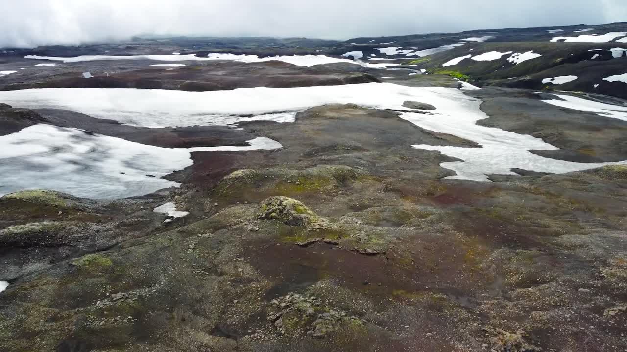 Aerial drone footage flying forward above Iceland dark brown and white snow covered volcanic rough terrain landscapes during a cloudy day with mist. Large dark mountains visible in the background.