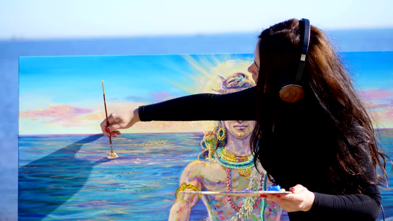여름, 강 근처, 해변. 해가 뜨면, 큰 갈색 헤드폰을 입은 아름다운 여성 예술가는 을 들고, 손에 팔레트를 들고, 캔버스 신 시바에 그림을 그리고, 음악을 듣고, 휴식을 취합니다.