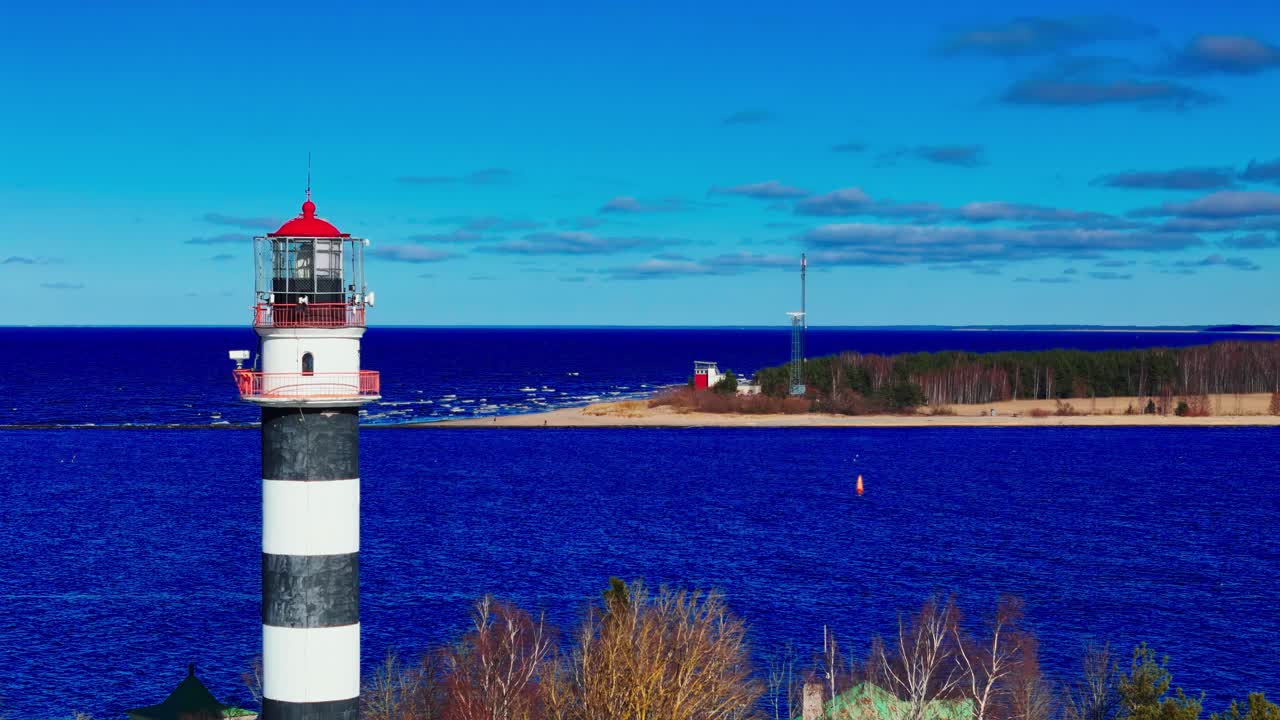 A striking black-and-white lighthouse stands tall above deep blue waves, framed by leafless trees and a pale sandy coast stretching to the horizon.
