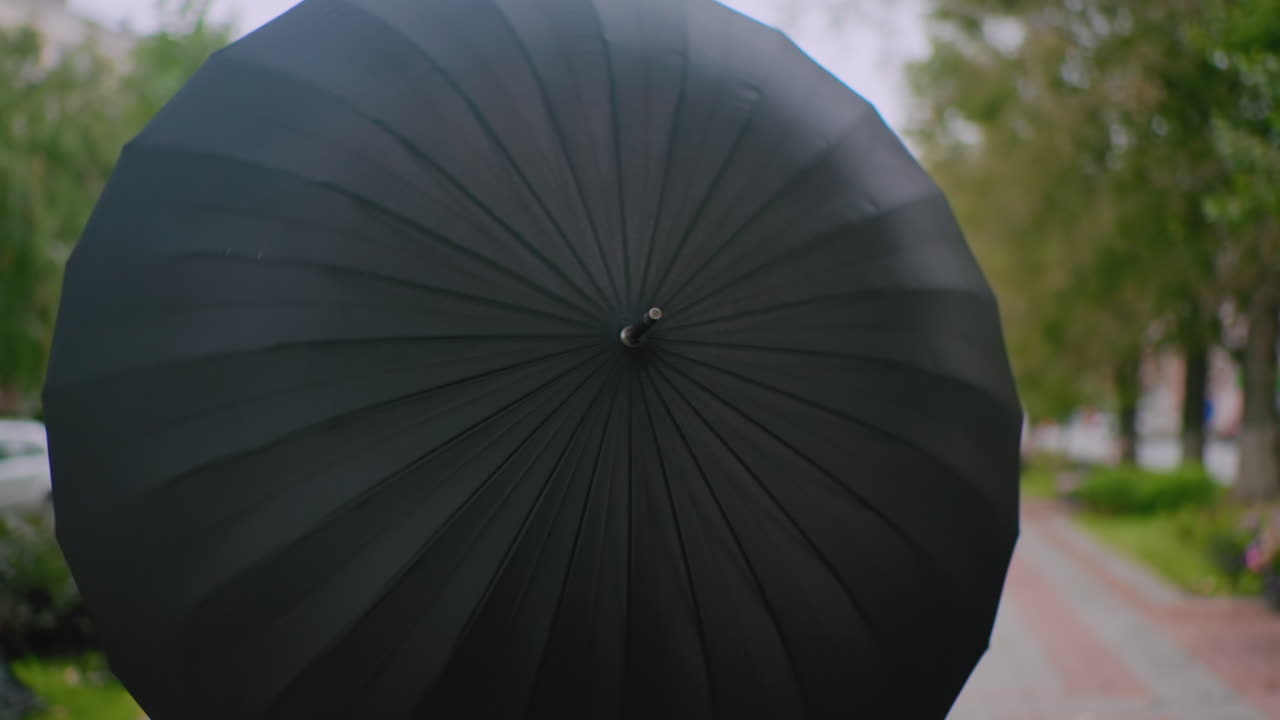 흐린 날, 도시의 거리에서 검은색 코트를 입고 검은색 우산을 펼쳐 돌리며 부드럽게 미소짓는 긴 머리의 젊은 여성이 나무, 사람들, 건물로 흐릿해진 배경 앞에 서 있어요.