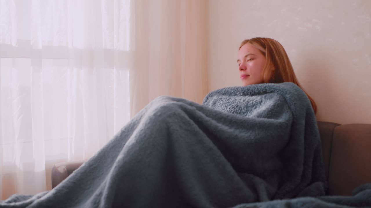 Lazy girl wrapped under fluffy blanket resting on couch near window with soft daylight entering through curtains, creating calm cozy atmosphere of relaxation comfort warmth