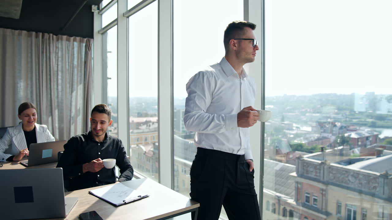 Positive young colleagues staying in office during lunch break. Fit man in white shirt stands at window with a cup in his hand looking at the cityscape.