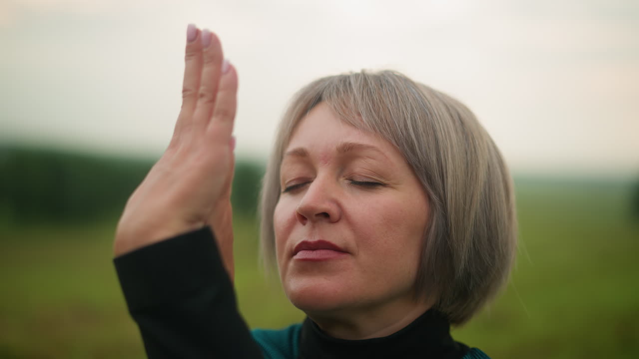 Close-up of woman in green and black suit in cow face pose, practicing yoga outdoors in a misty grassy field, hands raised in a meditative gesture with a soft blurred background