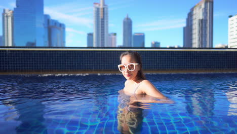 a young woman in a private rooftop resort pool turns from side to side against an urban skyline