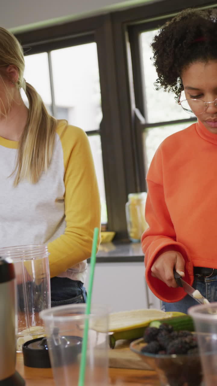 Vertical video of happy diverse teenager girls preparing healthy drink at home, slow motion