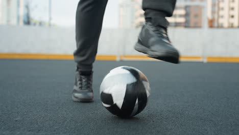 leg view of athlete in hoodie skillfully training with football on sport arena, tapping ball forward with precision, background includes chain-like fence and tall residential building