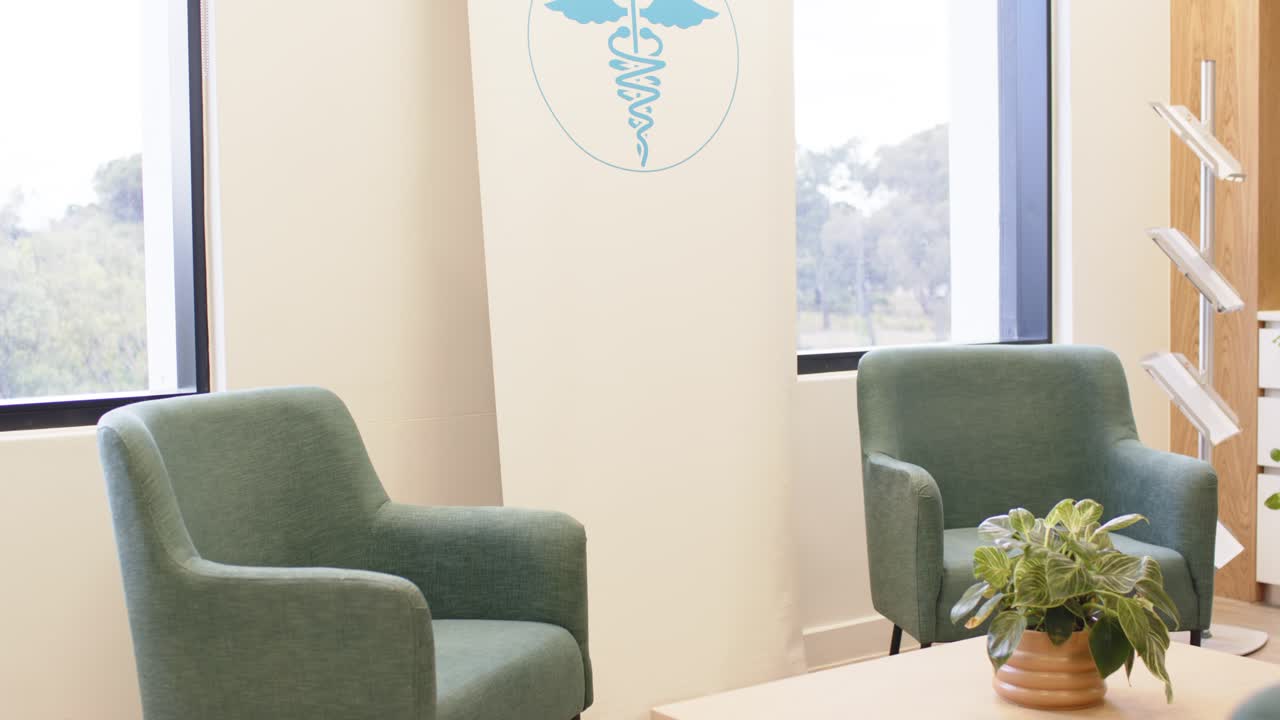 Camera kantelt en zwenkt, onthult de wachtruimte van de kliniek met een tafel, plantenpot en een banner die een caduceus omkadert.