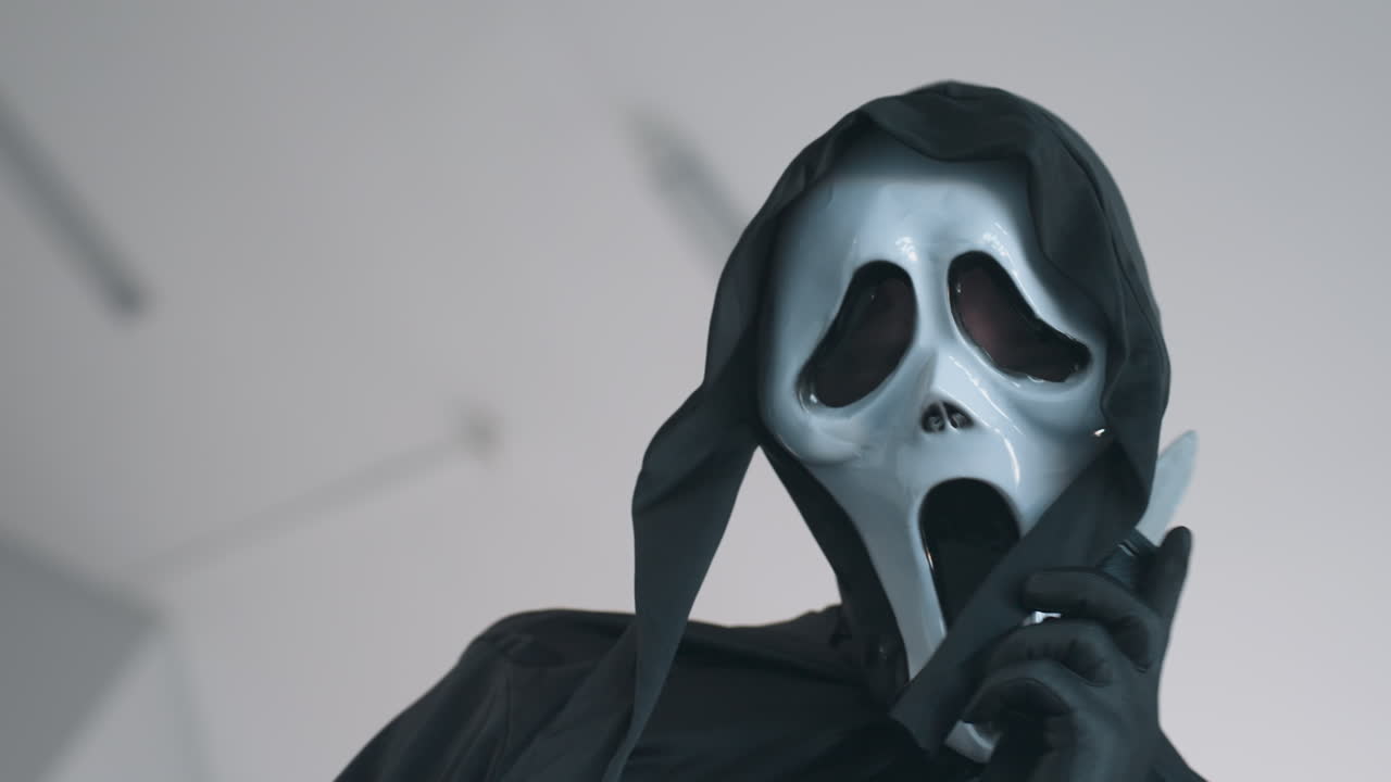 person in black cloak and white ghost mask holds knife near face in threatening gesture, wearing gloves in stark indoor setting with dramatic atmosphere and unsettling expression