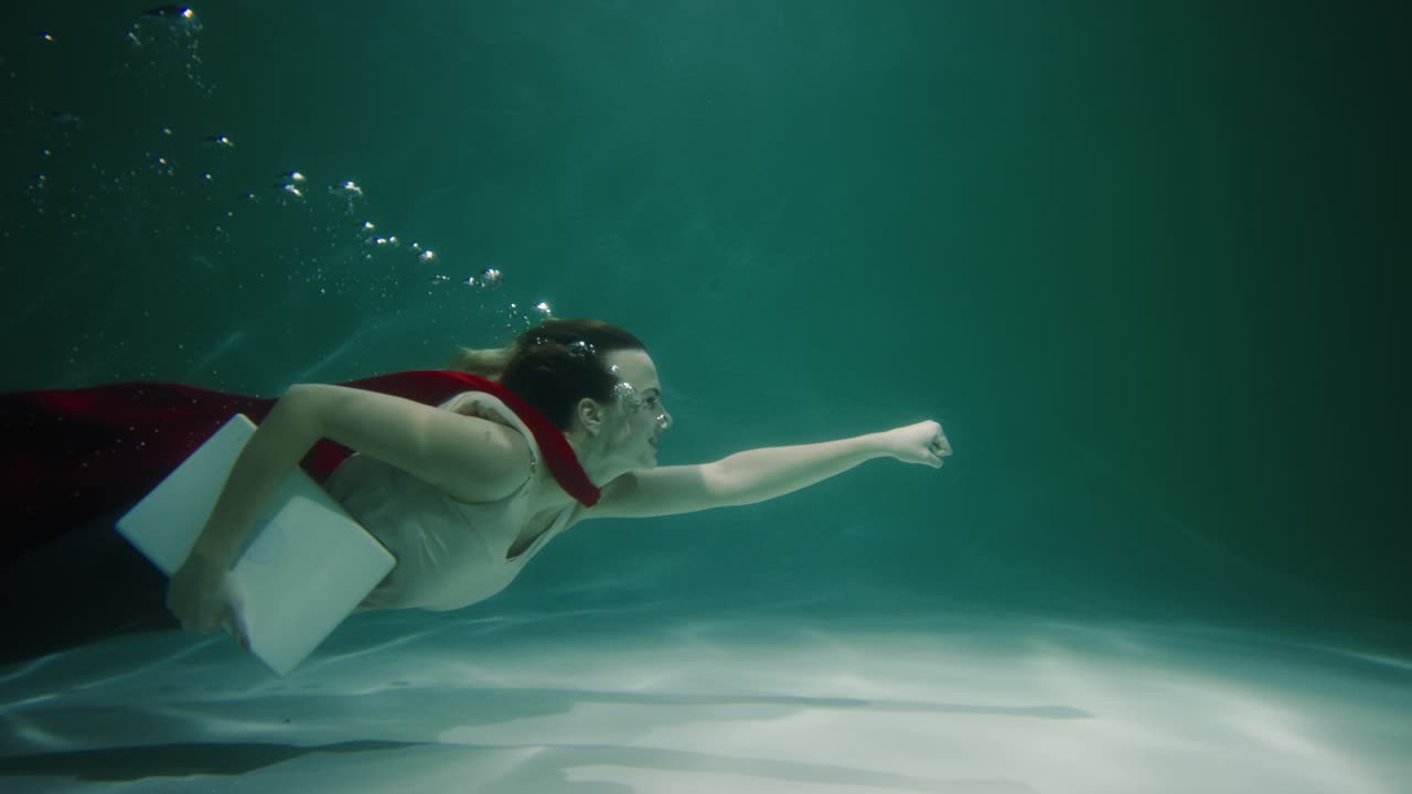 Rushing to help. Beautiful young female client service worker with red superhero cape suit swims underwater slow motion.