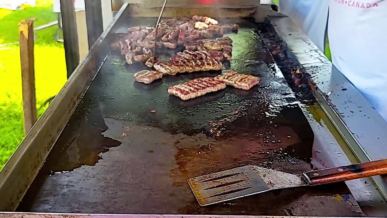 Primo piano di salsicce per colazione alla griglia durante una festa estiva all'aperto, con uno chef in uniforme bianca che separa la carne su una piastra usando una spatola, su uno sfondo di erba verde e soleggiata in una giornata calda e luminosa