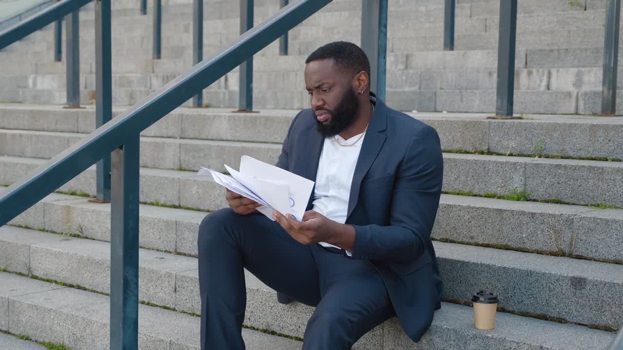 Bearded African American man looks through paper documents with business charts while sitting on the steps near the office center.
